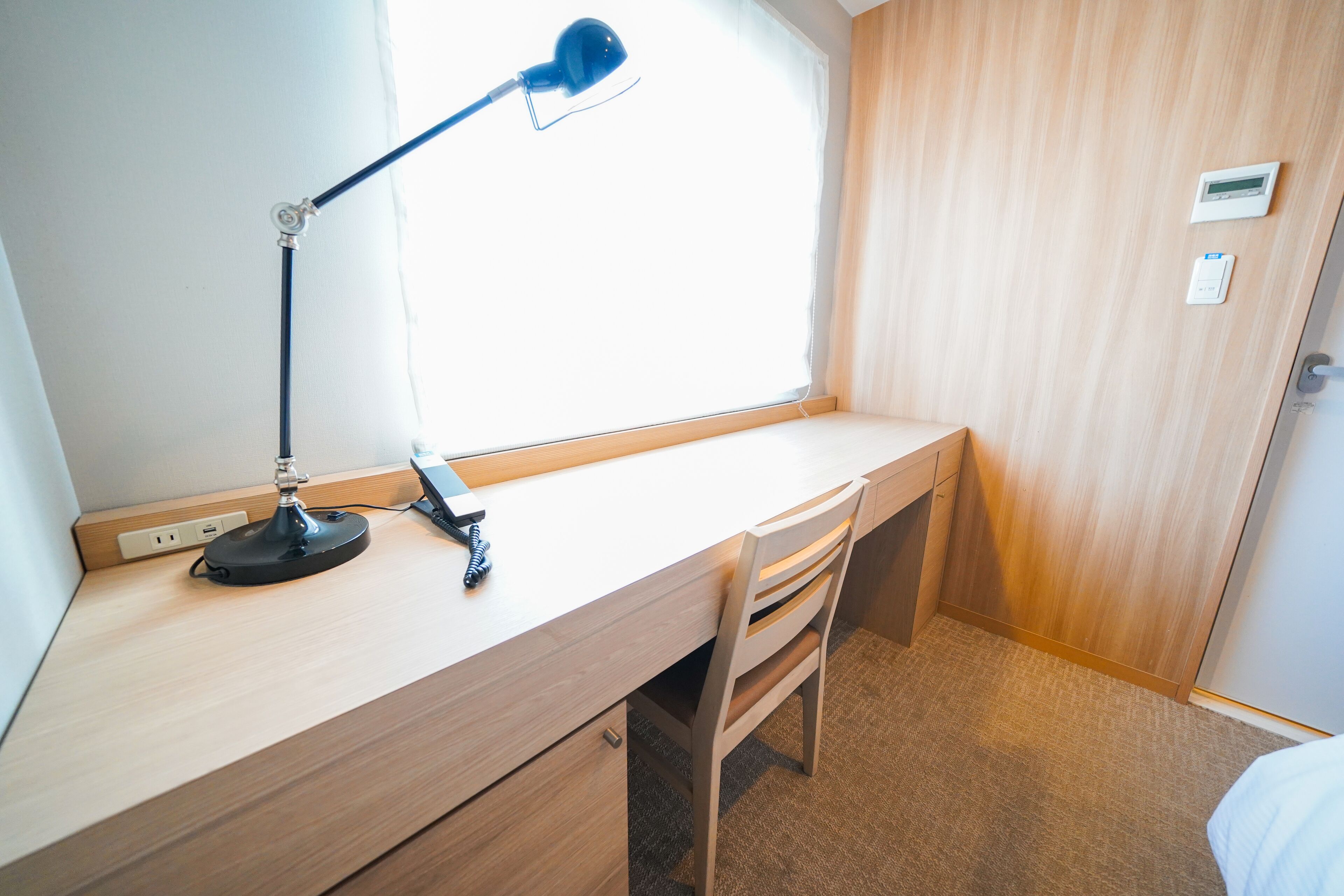 A room with a wooden desk, a blue desk lamp, a chair, a window with curtains, and a door with a thermostat.