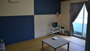 A room with a tatami mat floor, a small table with a book and remote, a white desk with a computer, a telephone, and a window with curtains. - Auberge ALBIREO (Toyooka)