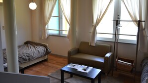 A small, well-lit room with a bed, a sofa, a table with a remote control, and a window with curtains. - Auberge ALBIREO (Toyooka)