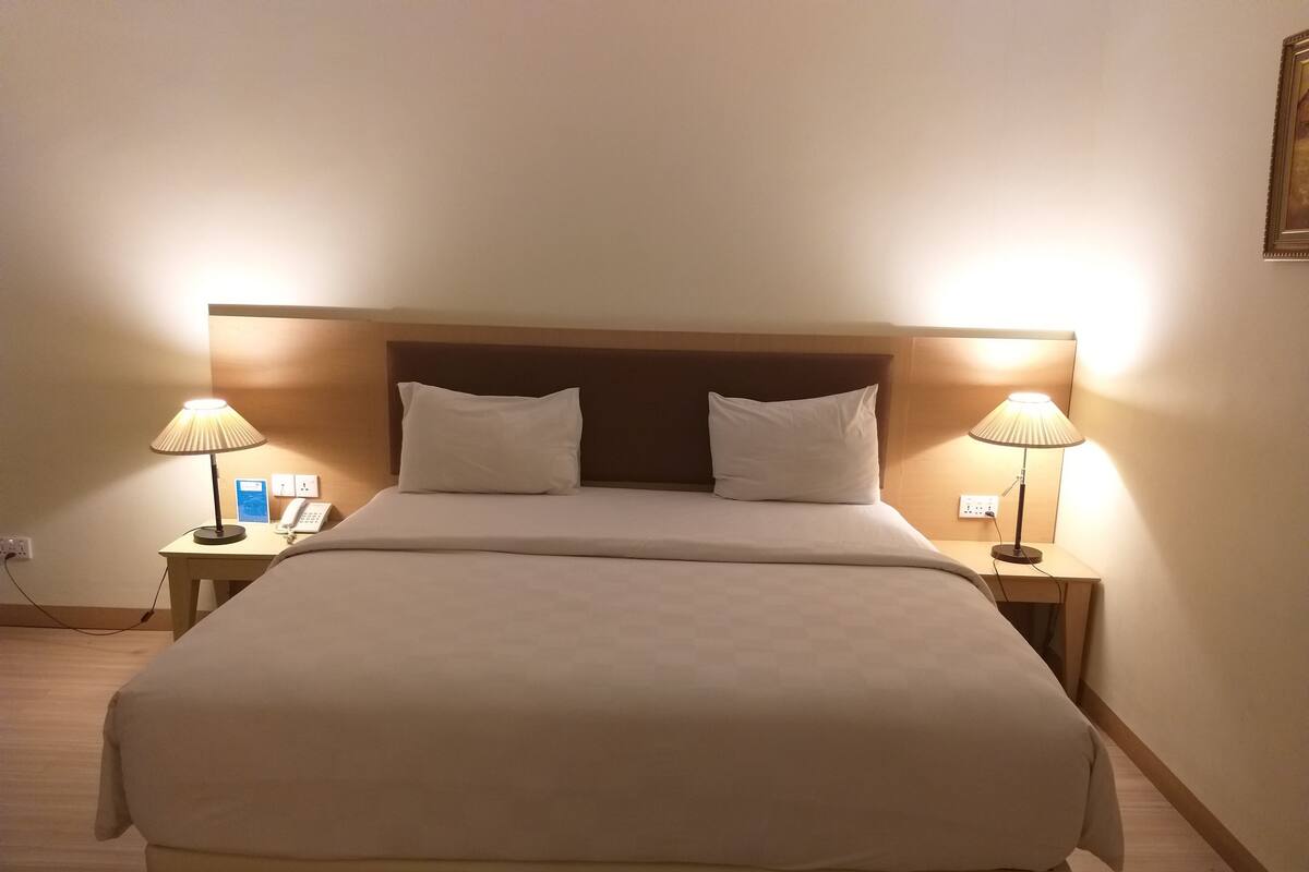 A neatly made bed with two pillows, flanked by two bedside tables with lamps, in a room with a simple wooden headboard and plain walls.