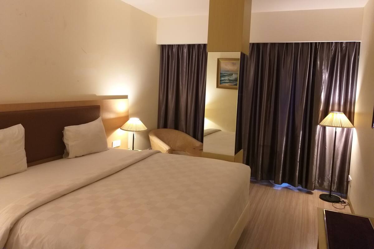A hotel room with a large bed, two bedside lamps, a chair, a mirror, and dark curtains.