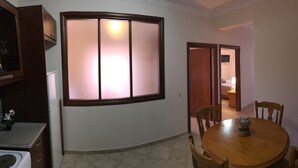 Habitació amb taula de dinar i cadires, una finestra gran amb panells de vidre rugós, i zona de cuina amb armaris i rentadora.