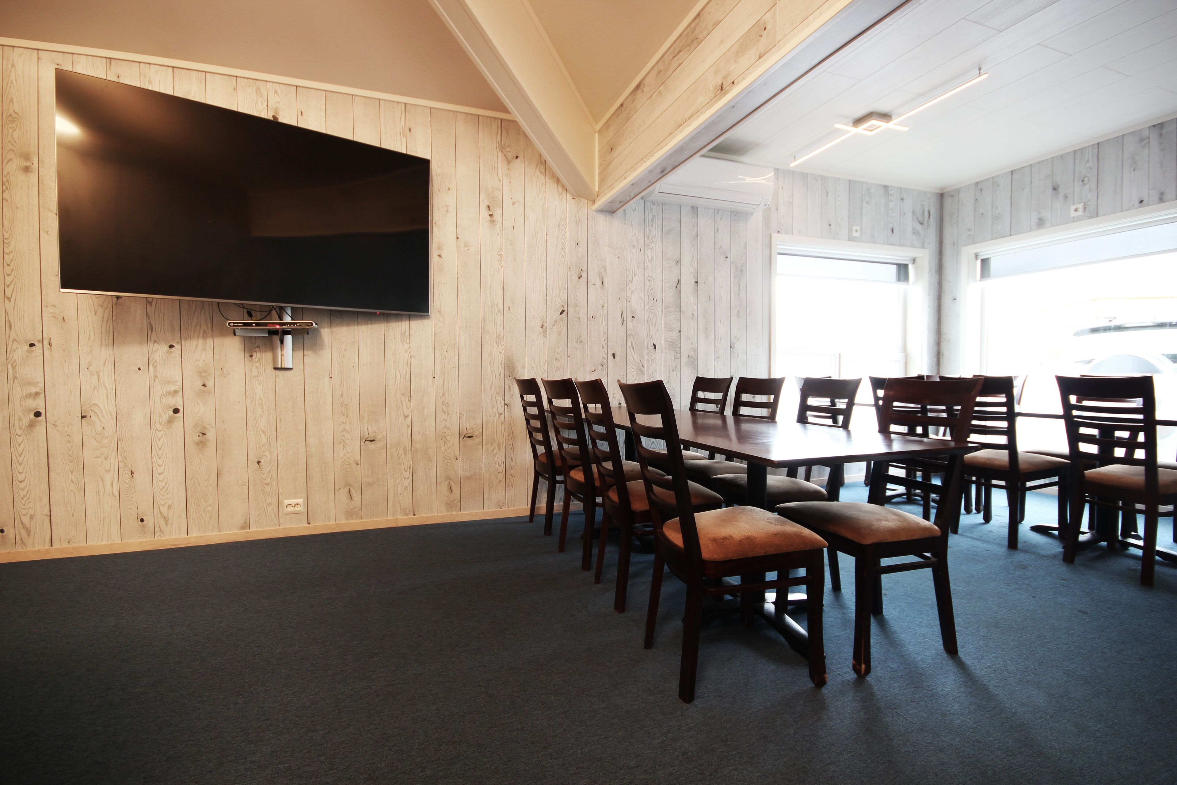 Une salle de conférence dotée d’un grand téléviseur à écran plat fixé au mur, de murs lambrés en bois, d’un sol recouvert d’une moquette sombre et d’une grande table entourée de chaises.