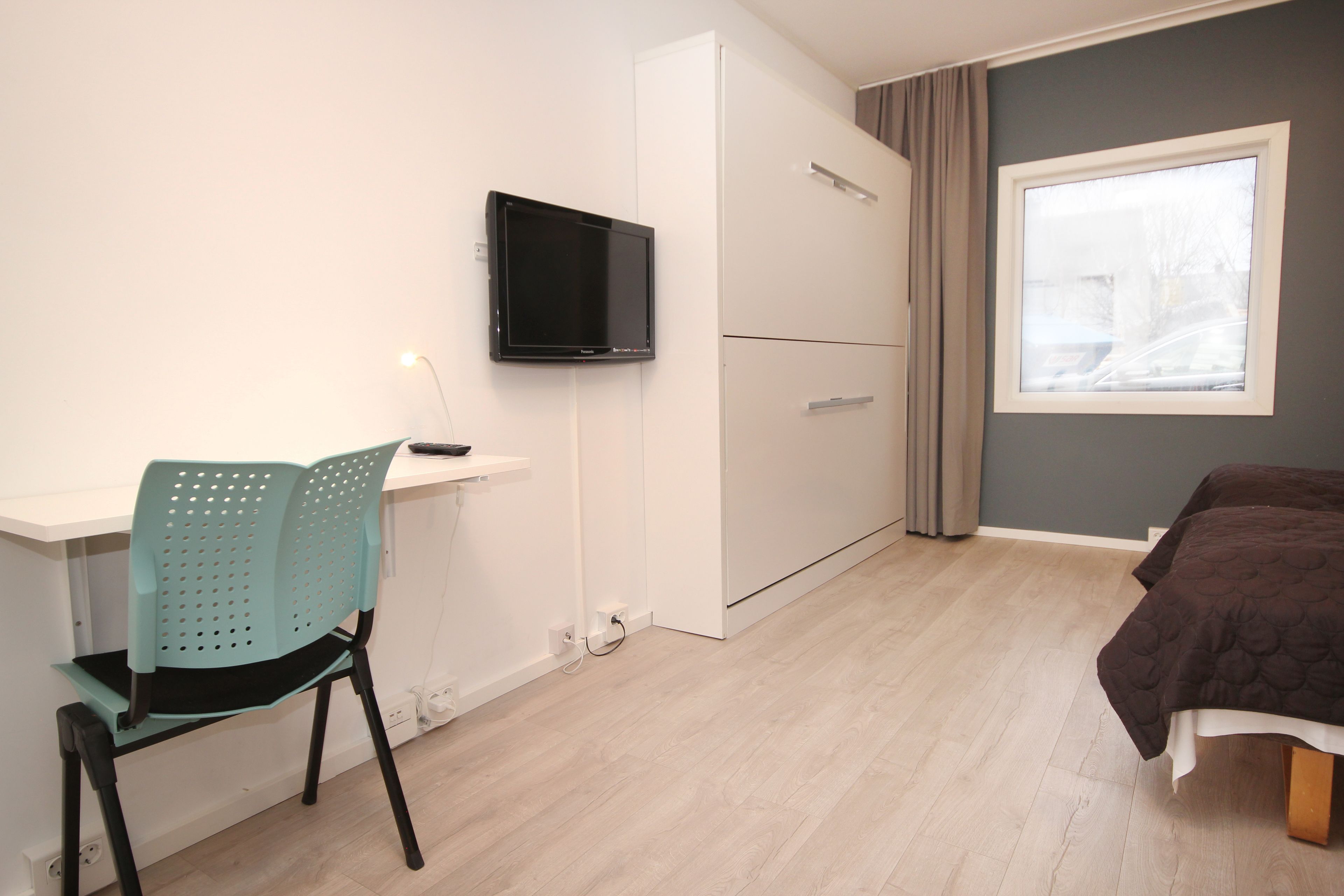 A bedroom with a bed, a desk with a chair, a TV, and a window with curtains.