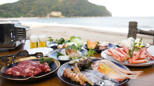 Uma mesa com diversos pratos de frutos do mar, uma churrasqueira e vista para a praia e oceano.