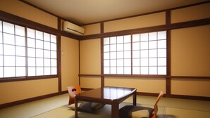 Um quarto com tatames, uma mesa de madeira baixa e dois almofados, janelas amplas com molduras de madeira, e um ar-condicionado fixado na parede.