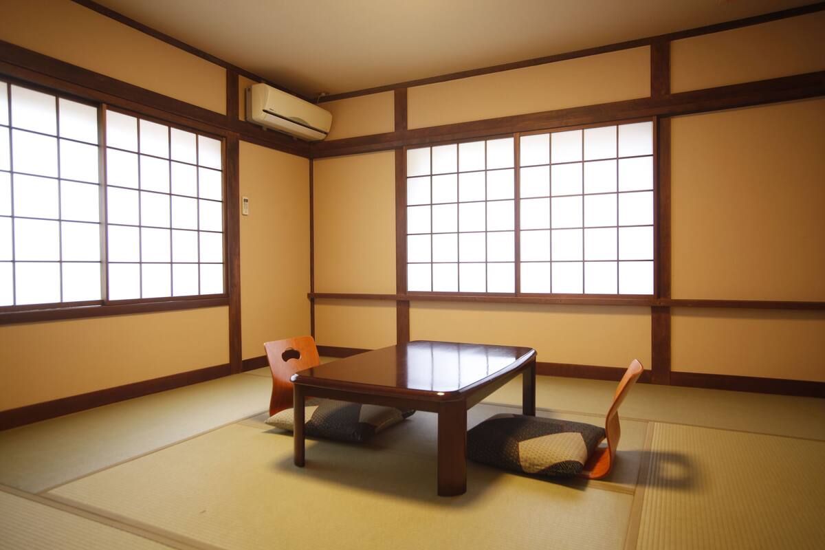 A tatami room with a low wooden table and two cushions, large windows with wooden frames, and a wall-mounted air conditioner.