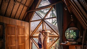 A cozy bedroom with a large window, wooden furniture, and a bed with a patterned bedspread. - Gili Treehouses (Gili Trawangan)