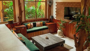 A cozy living room with a sofa, coffee table, and a TV. There are large windows with a view of greenery outside. - Gili Treehouses (Gili Trawangan)