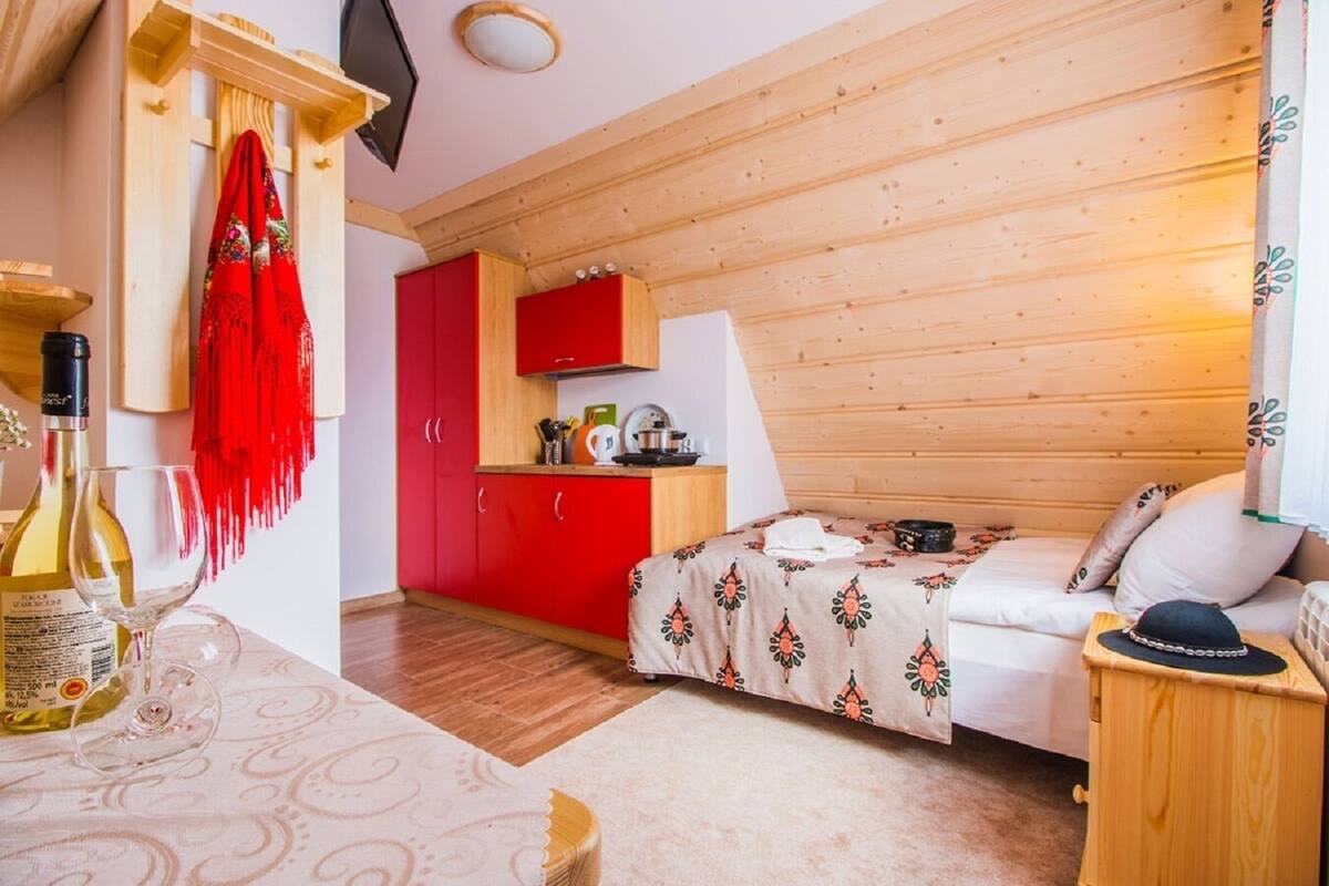 Una habitación acogedora con una cama, una mesita con una botella y un vaso, un armario rojo y un techo de madera.