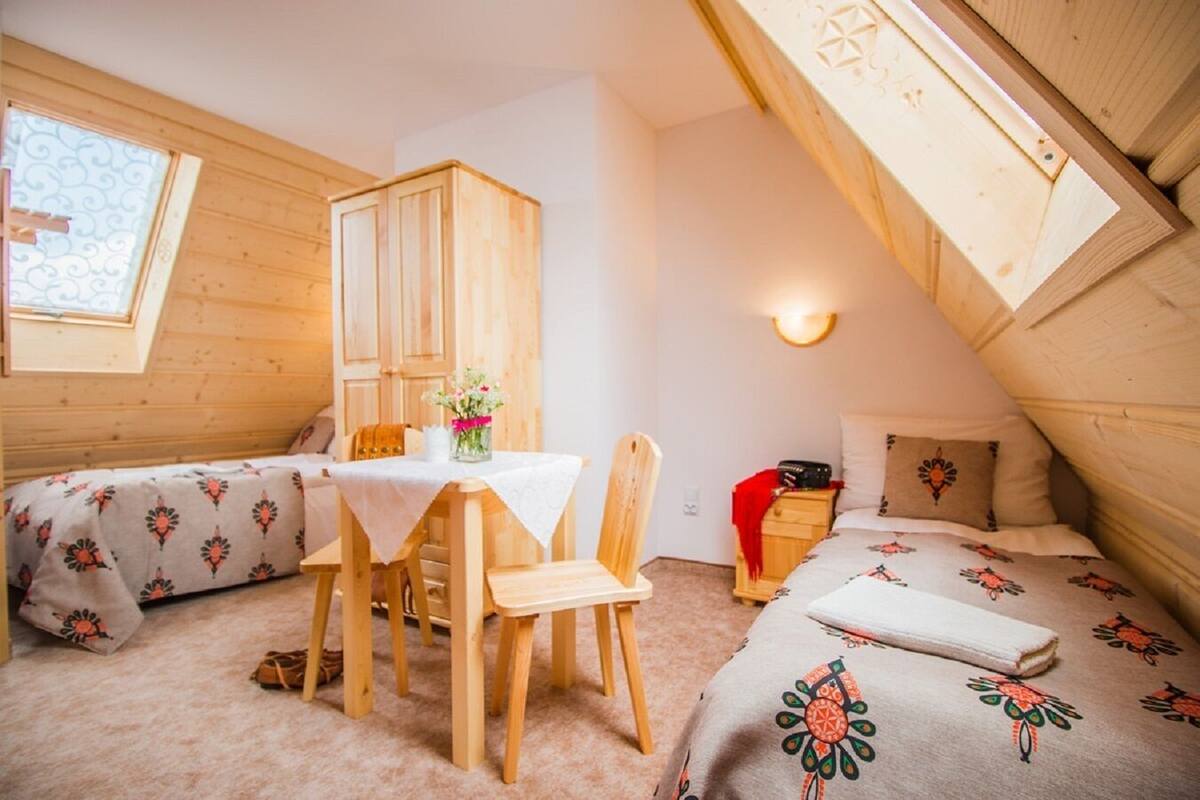 Un dormitorio acogedor en el ático con techo de madera, una mesa de comedor con sillas, una cama con ropa de cama floral y una mesita de noche pequeña con una lámpara.