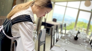 Una persona preparando una mesa con copas de vino y una botella, dentro de una estructura similar a una carpa, con vistas a zonas verdes.