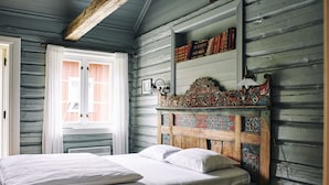Un dormitorio con cabecera de madera, cama con ropa blanca, ventana con cortinas blancas y estante con libros.