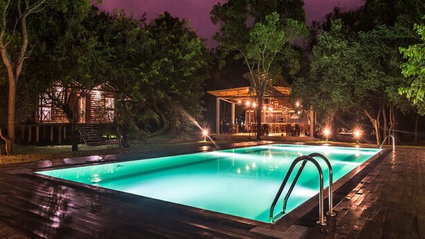 A swimming pool with a wooden deck, surrounded by trees and a building with outdoor seating. - Eighth Wonder Resort (Sigiriya)