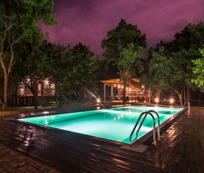 A swimming pool with a wooden deck, surrounded by trees and a building with outdoor seating. - Eighth Wonder Resort (Sigiriya)