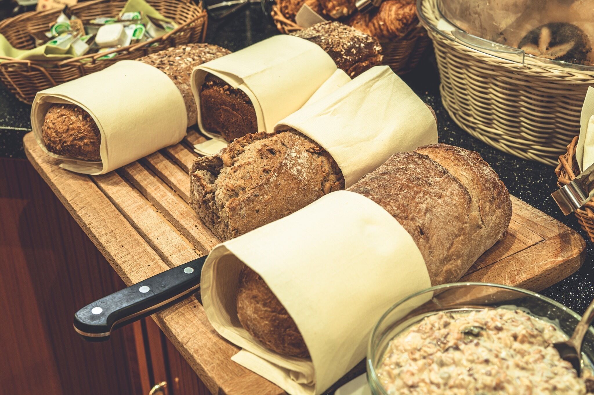 A wooden cutting board with several loaves of bread wrapped in paper, a knife, and a bowl of spread.