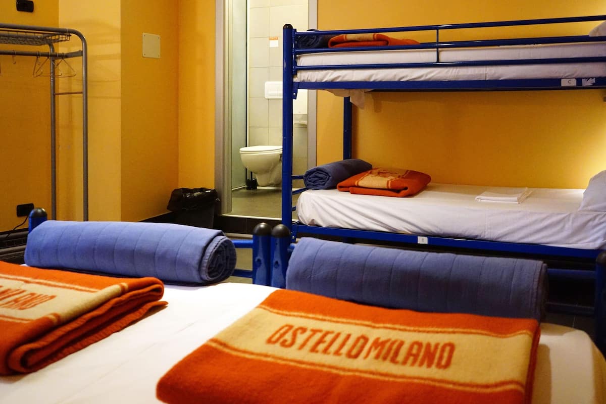 Une chambre avec des lits superposés, un présentoir en métal, des toilettes et des serviettes portant la marque « Ostello Milano ».