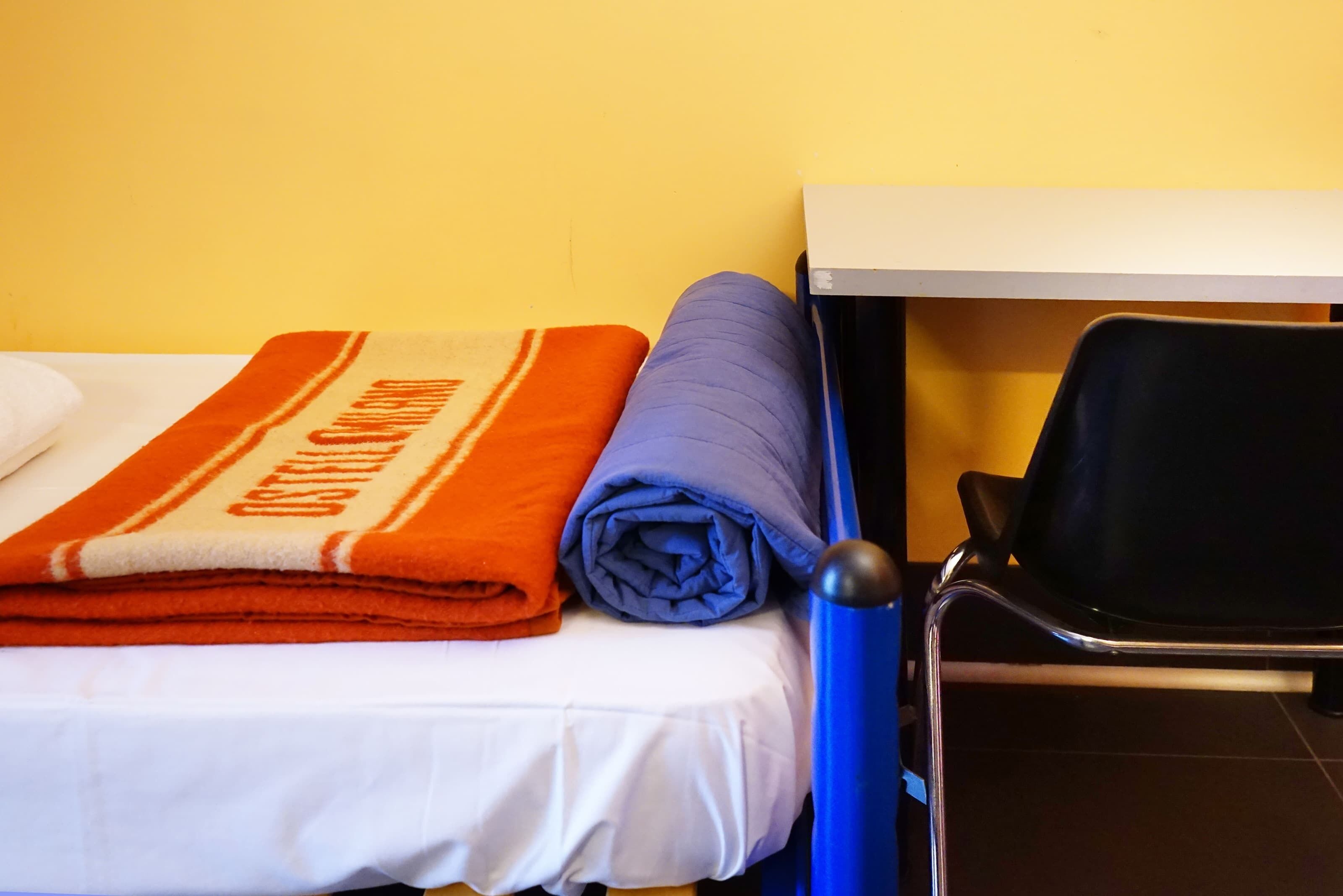A bed with a white sheet and a folded orange and white blanket, a blue pillow, and a black chair next to a white table.
