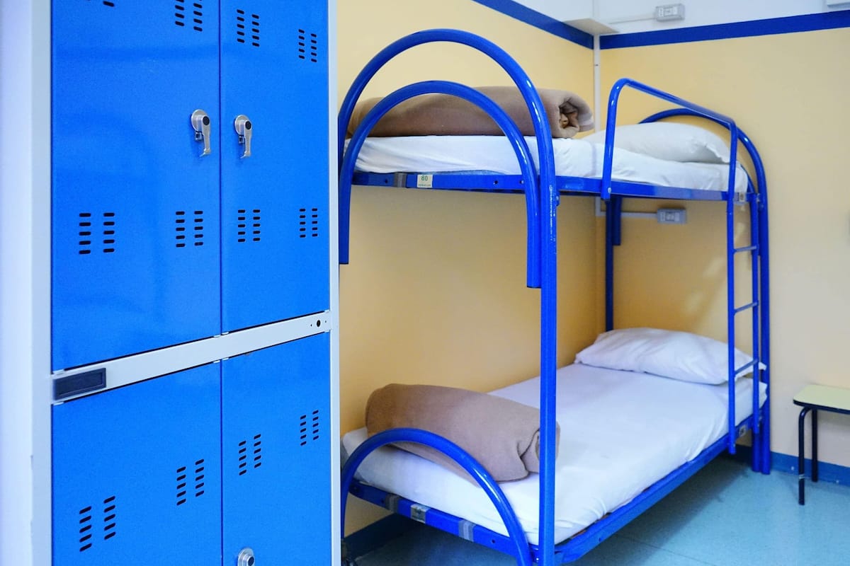 Un lit superposé avec une structure en métal bleue et un matelas recouvert d’une couverture marron. On y trouve des casiers bleus dotés d’orifices de ventilation, ainsi qu’une petite table placée à côté du lit.
