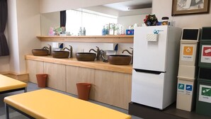 A modern kitchen with wooden cabinets, a white refrigerator, and a sink with multiple bowls. There are trash bins and a bench in the room. - Hostel Mallika (Hiroshima)
