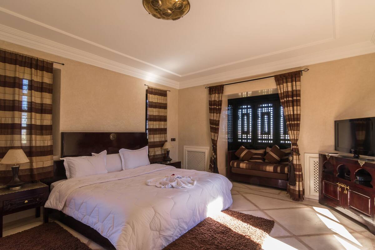 Quarto de hotel com uma cama grande, cabeceira de madeira, televisão, sofá e cortinas.