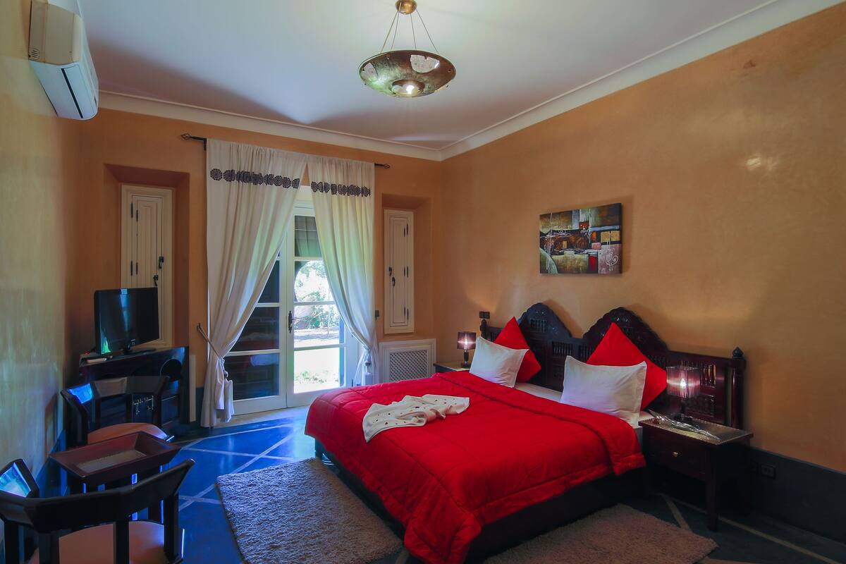 Quarto com colcha vermelha, televisão em suporte, escrivaninha e uma cadeira.