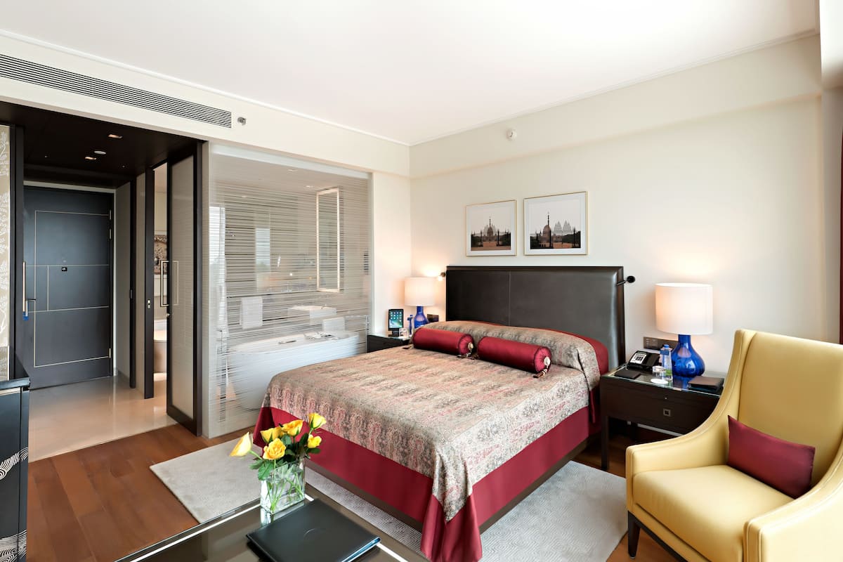 Moderni hotellihuone, jossa on suuri sänky, yöpöydät, tuoli ja ikkunasta avautuva näkymä kaupunkiin.