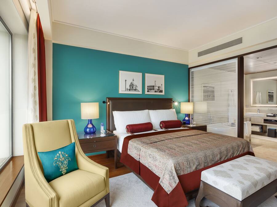 A hotel room with a large bed, two bedside tables, a chair, a window with curtains, and a bathroom visible through a glass door.