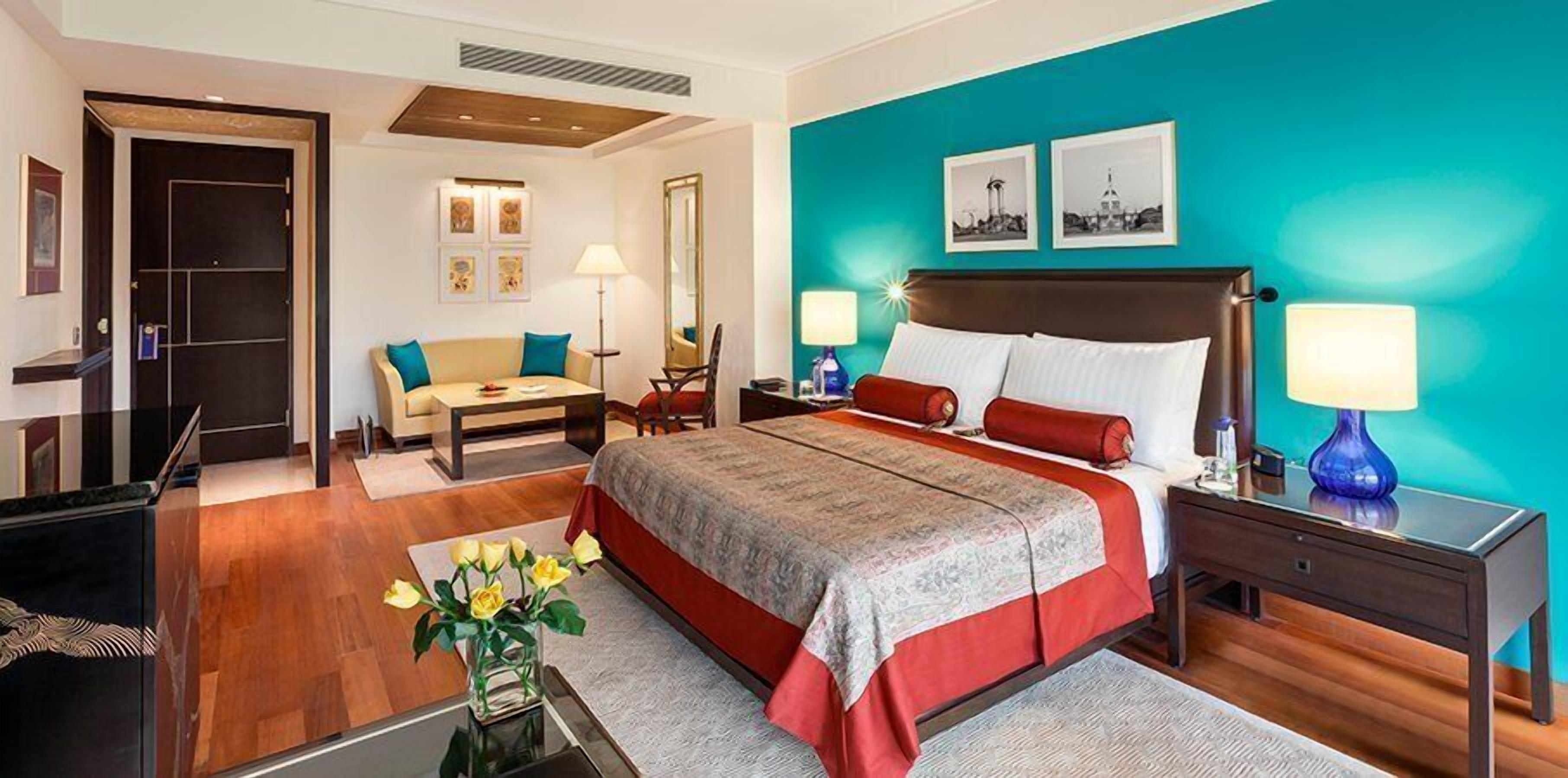 A hotel room with a large bed, a desk, a chair, a sofa, and a coffee table. There are two lamps on the nightstands and a vase with flowers on the coffee table.