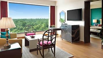 A hotel room with a large window offering a view of a forest, a flat-screen TV mounted on the wall, a desk with a lamp, and a bed with a nightstand.