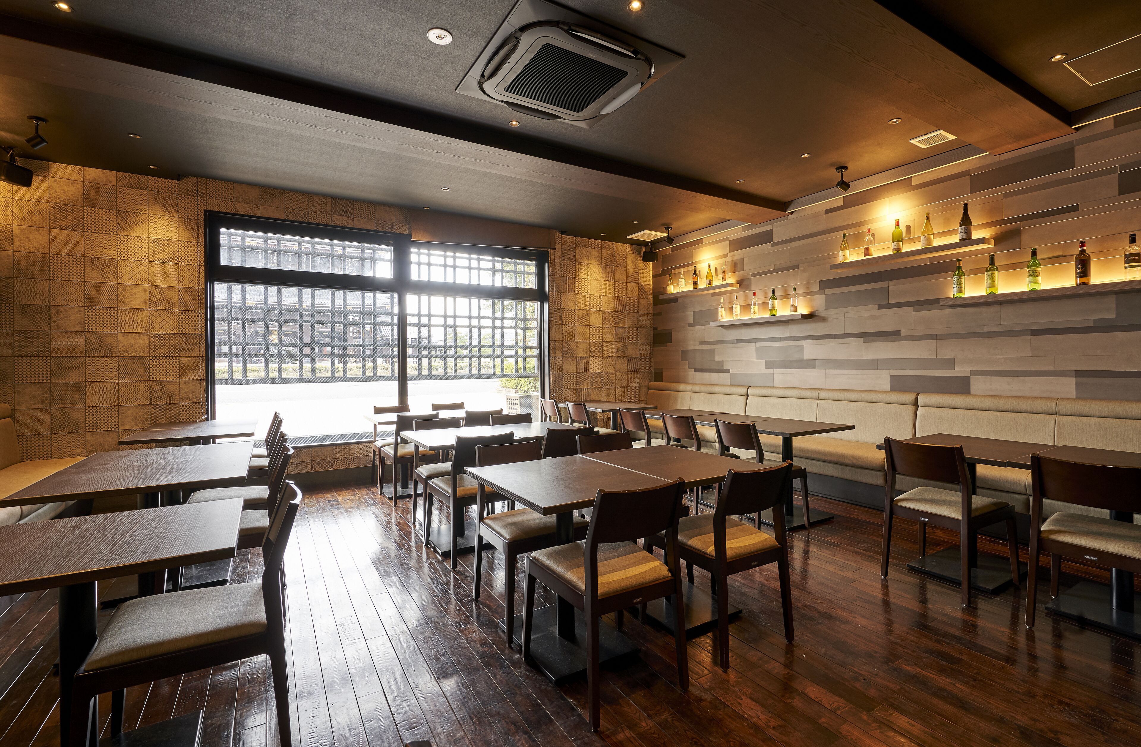A restaurant interior with wooden tables and chairs, a brick wall with shelves of bottles, and large windows.