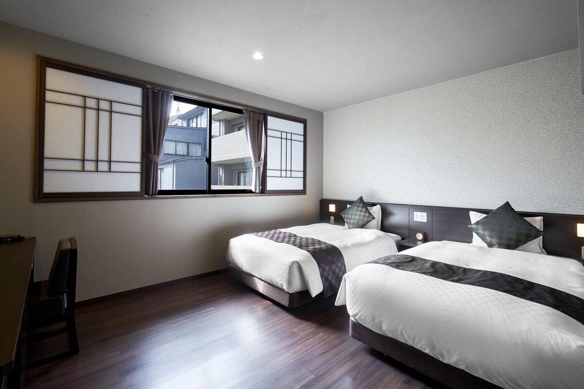 Habitación de hotel con dos camas, piso de madera y un ventanal con vistas a edificios.