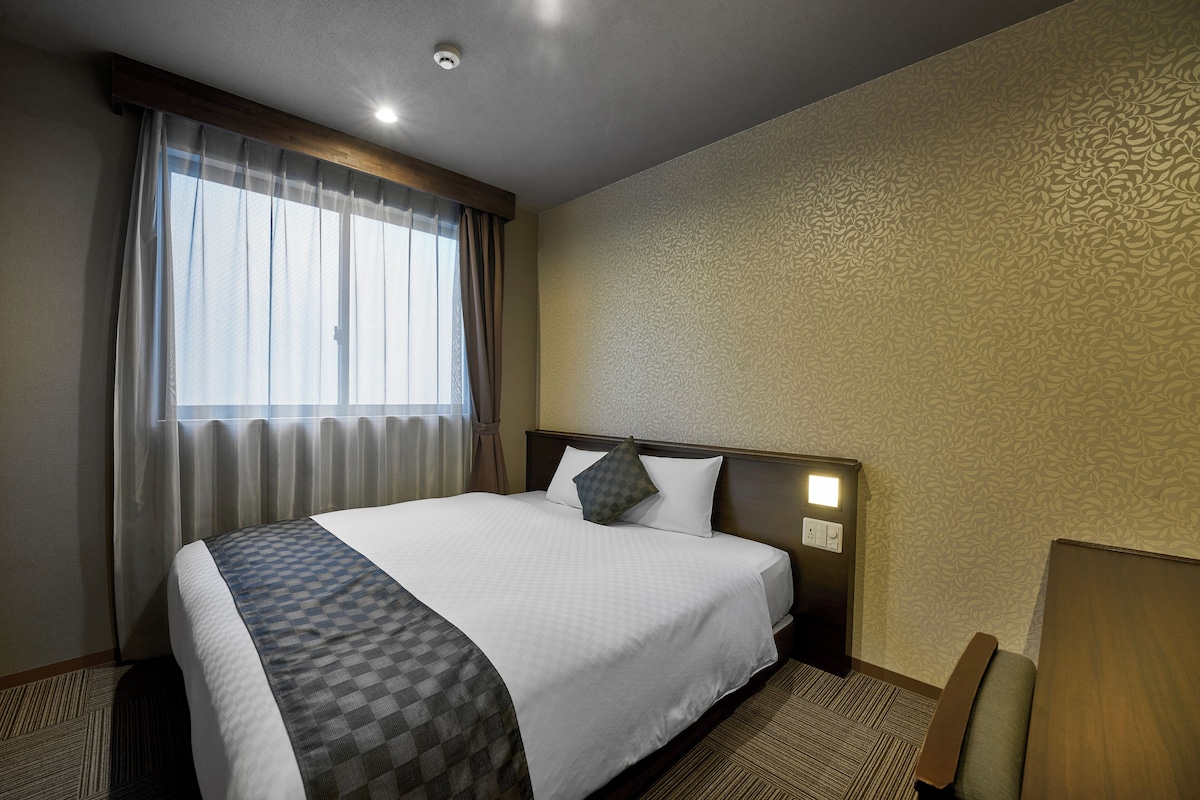 Habitación de hotel con una cama grande, cabecera con lámpara, mesita de noche, una silla y una ventana con cortinas.