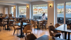 Un restaurante moderno con mesas y sillas, amplios ventanales con vista al paseo marítimo y edificios.
