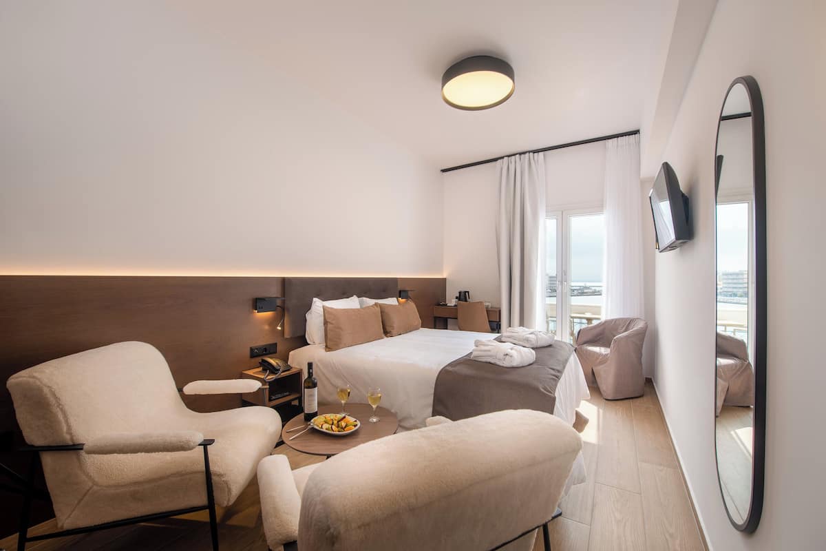 A modern hotel room with a bed, two armchairs, a small table with a bottle and glasses, and a large mirror.