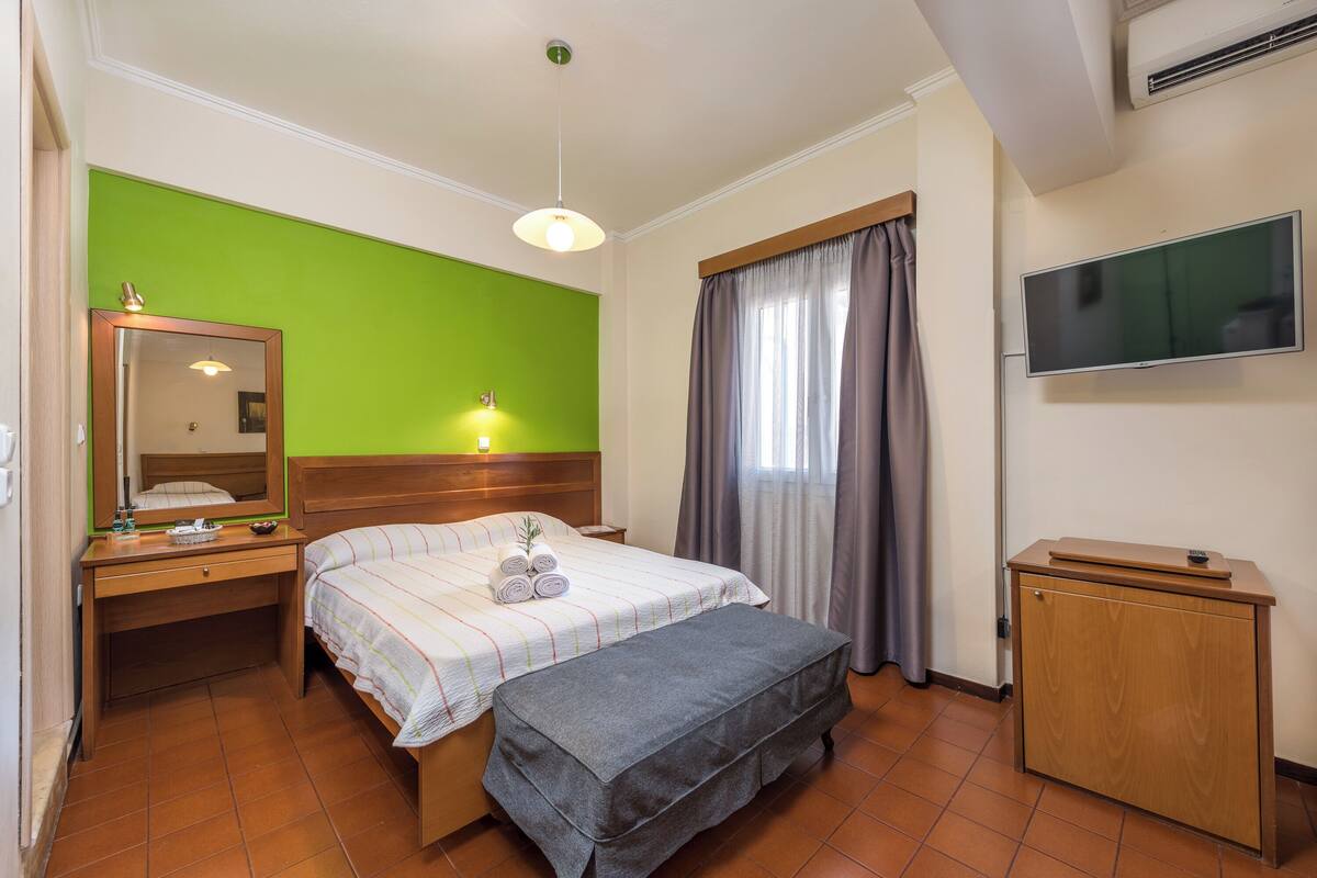 A hotel room with a bed, bedside table, mirror, lamp, TV, and a small wooden cabinet.
