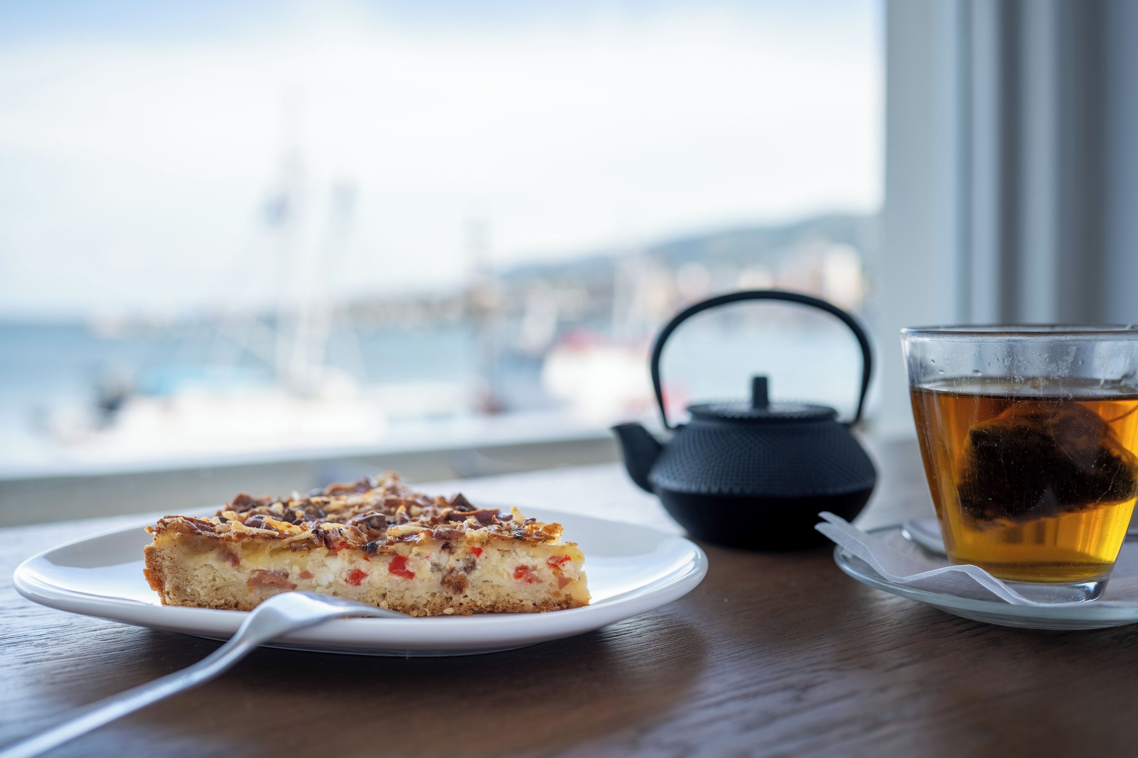 A slice of cake on a plate, a cup of tea with a teapot, and a view of a body of water through a window.