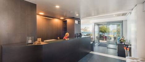 Un área de recepción moderna con un mostrador oscuro, una recepcionista y una puerta de cristal que da al exterior.