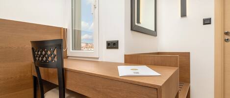 A wooden desk with a chair, a window with a view of buildings, a mirror, and a door.