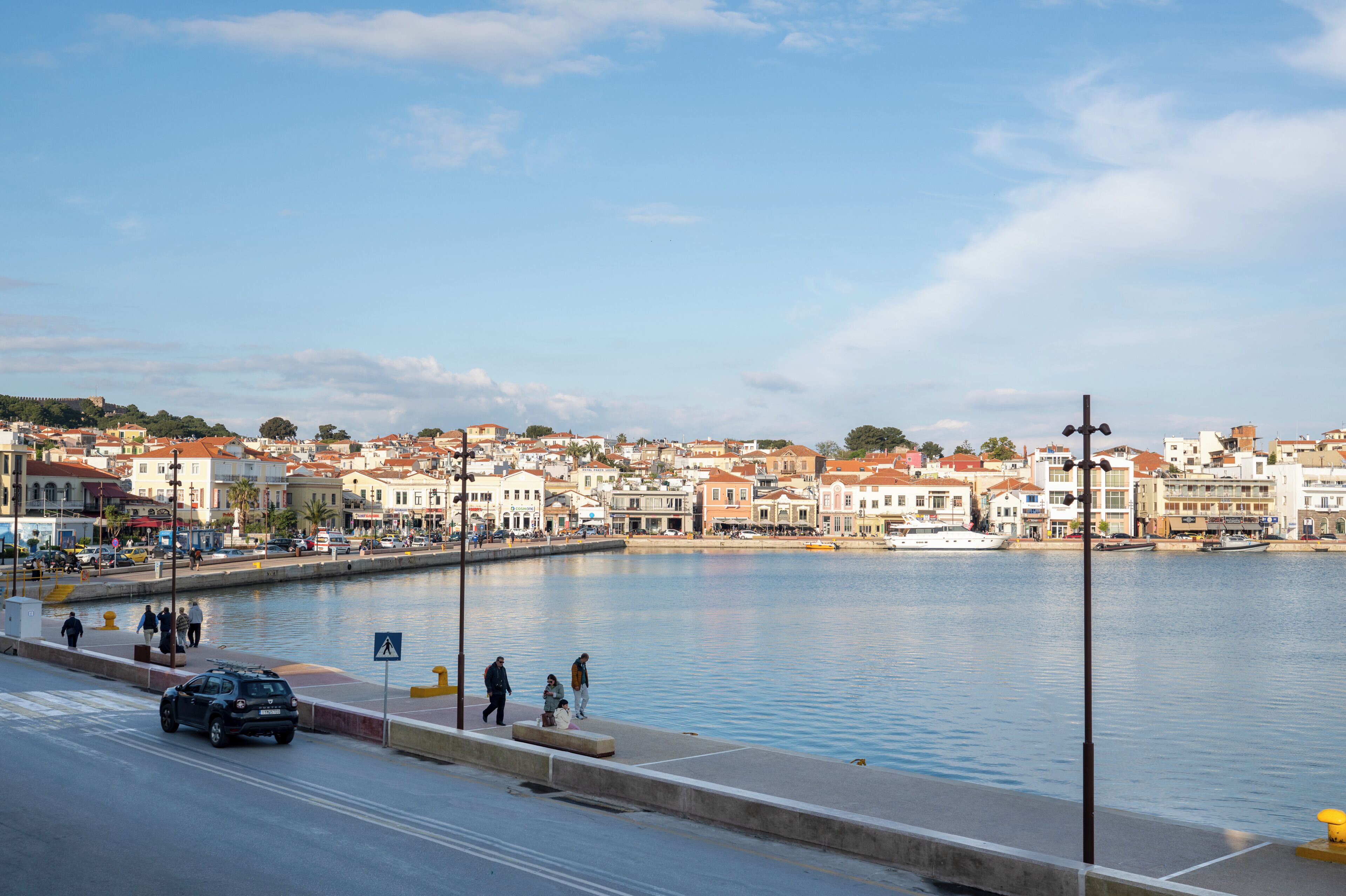 A waterfront cityscape with buildings, a calm river, and a pedestrian walkway.