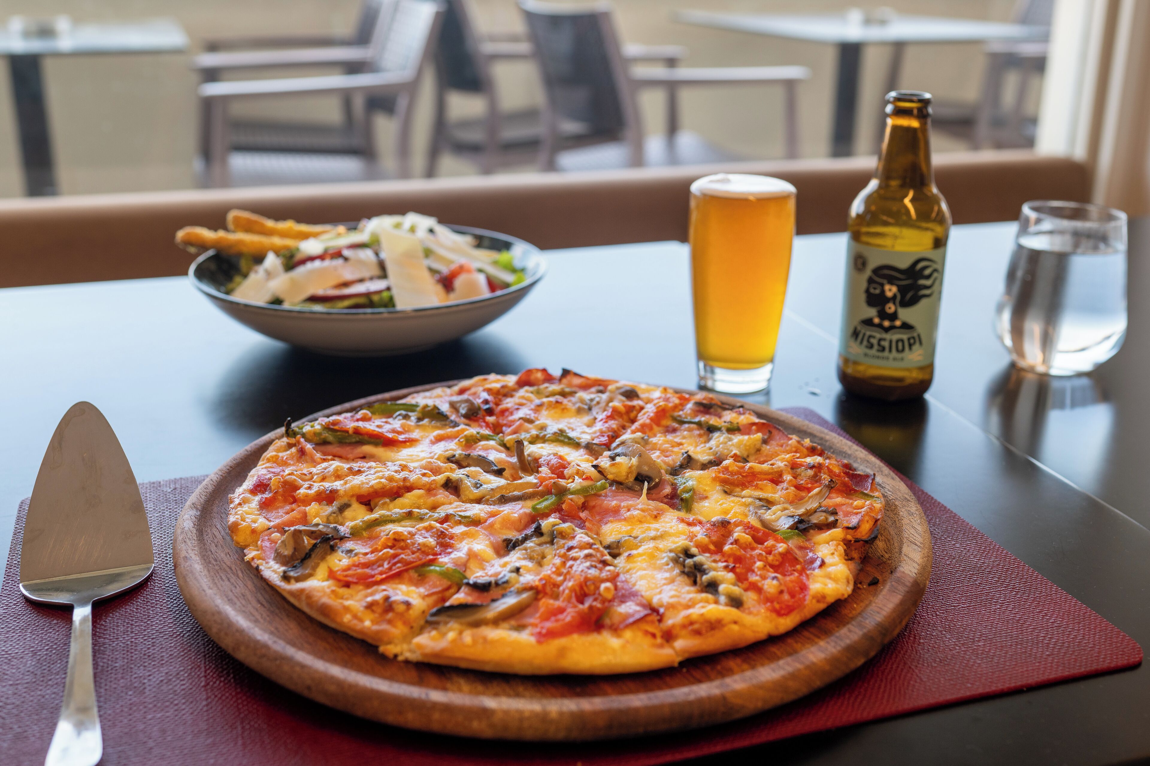 A pizza on a wooden board, a beer bottle, a glass of beer, and a bowl of salad on a table.