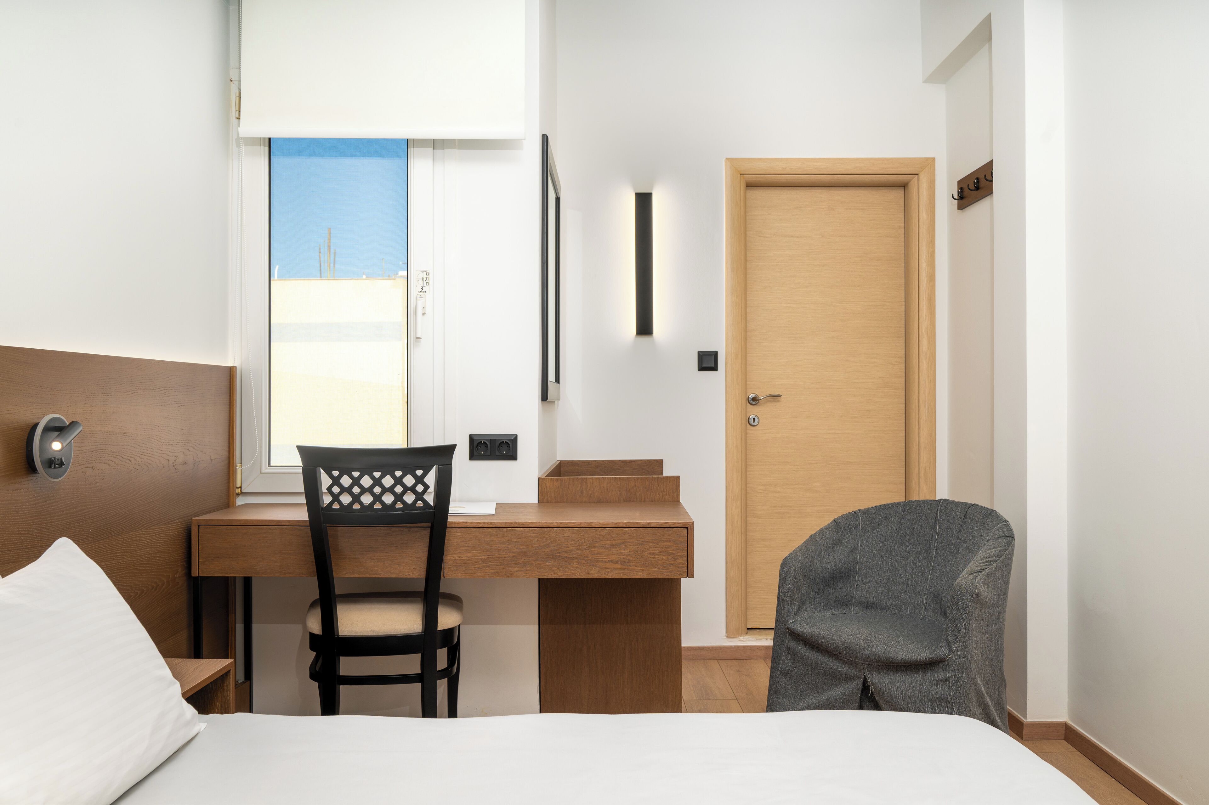 Habitación de hotel con cama, escritorio con silla, puerta de madera y ventana con vista a edificios.