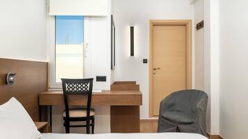 Habitación de hotel con cama, escritorio con silla, puerta de madera y ventana con vista a edificios.