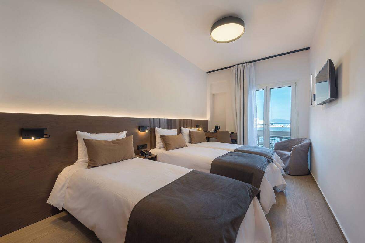 A hotel room with two beds, a TV, a bedside table, a lamp, and a window with curtains.