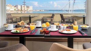 Una mesa de desayuno con varios platos, vista al mar y edificios al fondo.