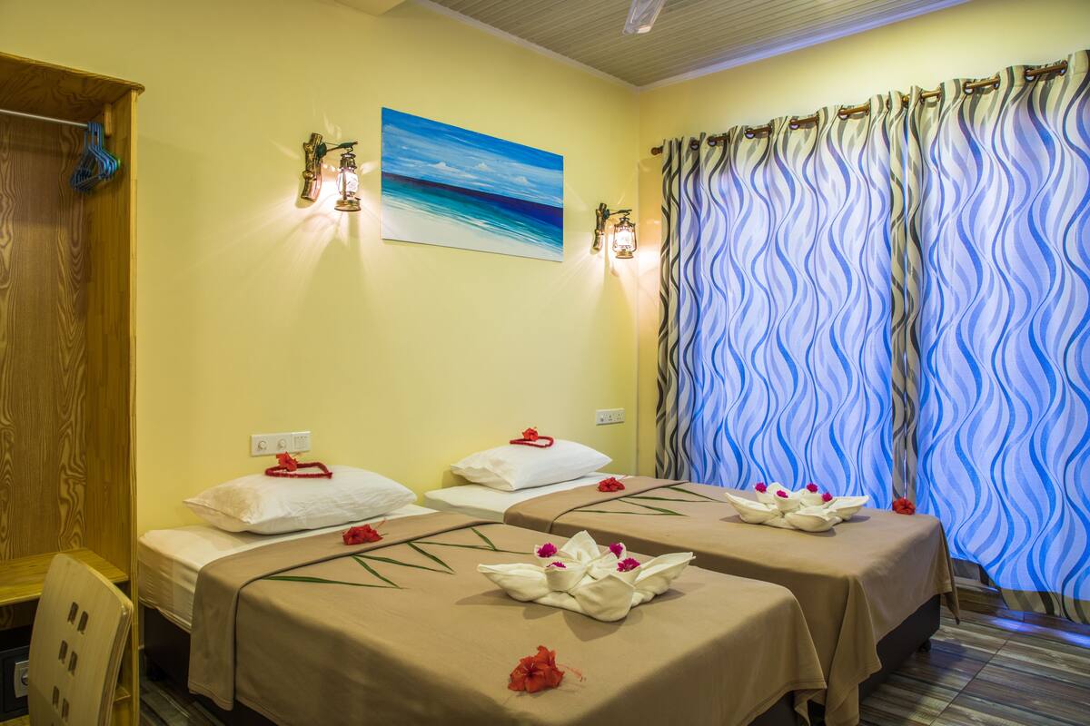 A room with two beds, each with a towel and a flower. There is a painting on the wall and a curtain with a wavy pattern.
