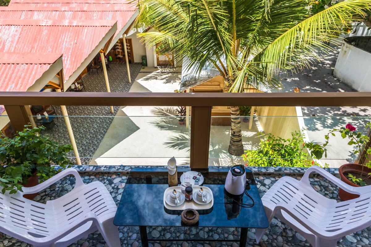 A balcony with a table set for two, two plastic chairs, a potted plant, and a view of a pool and palm trees.