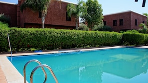 Una piscina con agua cristalina azul, rodeada por un seto cuidado y un edificio de ladrillo al fondo.