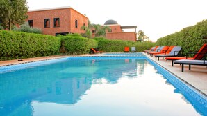 En swimmingpool med solstolar, en byggnad i tegel med en kupol och välskötta häckar.