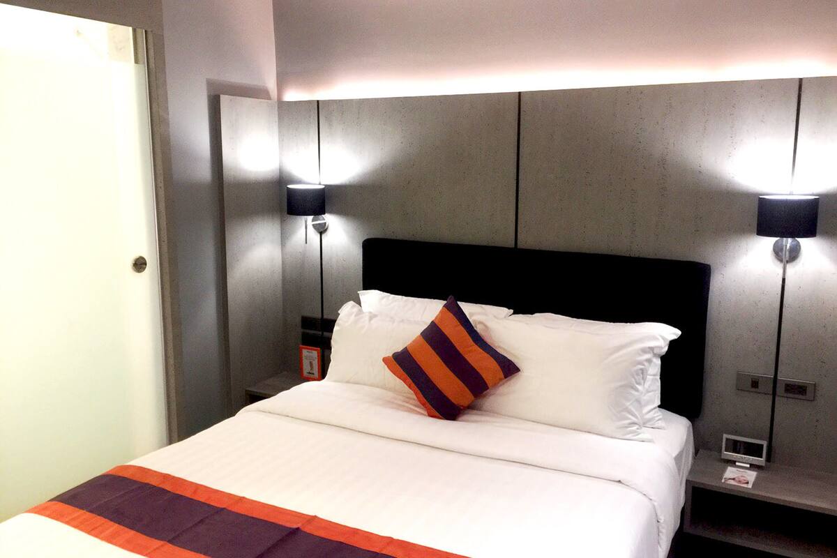 Una habitación de hotel con cama, mesita de noche, dos lámparas de pared y una puerta.